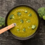 A bowl of creamy homemade broccoli soup garnished with small broccoli florets, surrounded by fresh broccoli heads on a rustic wooden background.