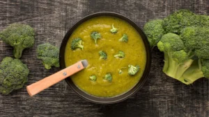 A bowl of creamy homemade broccoli soup garnished with small broccoli florets, surrounded by fresh broccoli heads on a rustic wooden background.