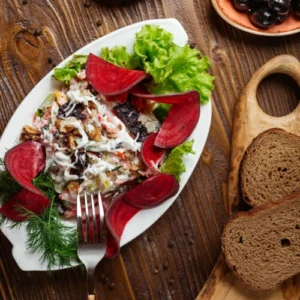 A vibrant beet salad served on a white plate with lettuce, fresh dill, sour cream drizzle, and slices of beetroot, placed on a rustic wooden table alongside rye bread and a fork.