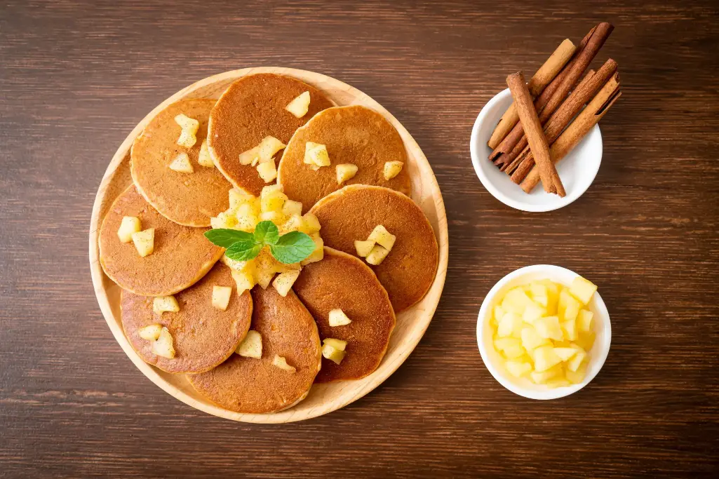 A plate of apple pancakes topped with diced apples and garnished with a sprig of mint, served alongside bowls of cinnamon sticks and diced apples.