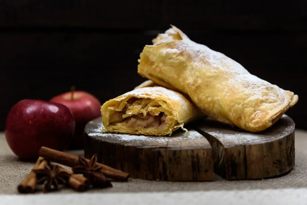 Freshly baked apple strudel dusted with powdered sugar, placed on a wooden board, surrounded by fresh apples, cinnamon sticks, and star anise.