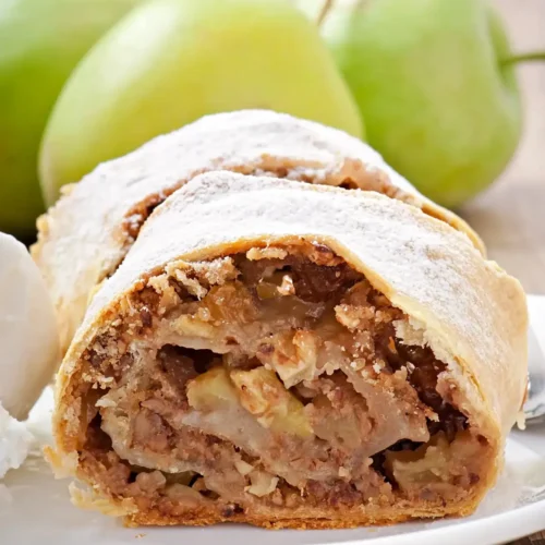 Close-up of a slice of apple strudel filled with spiced apples and raisins, dusted with powdered sugar, served on a plate with a scoop of vanilla ice cream, and green apples in the background.