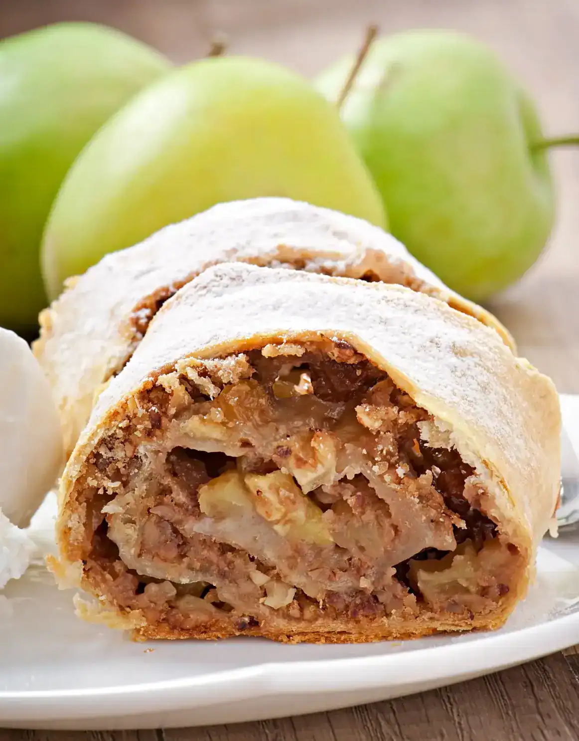 Close-up of a slice of apple strudel filled with spiced apples and raisins, dusted with powdered sugar, served on a plate with a scoop of vanilla ice cream, and green apples in the background.