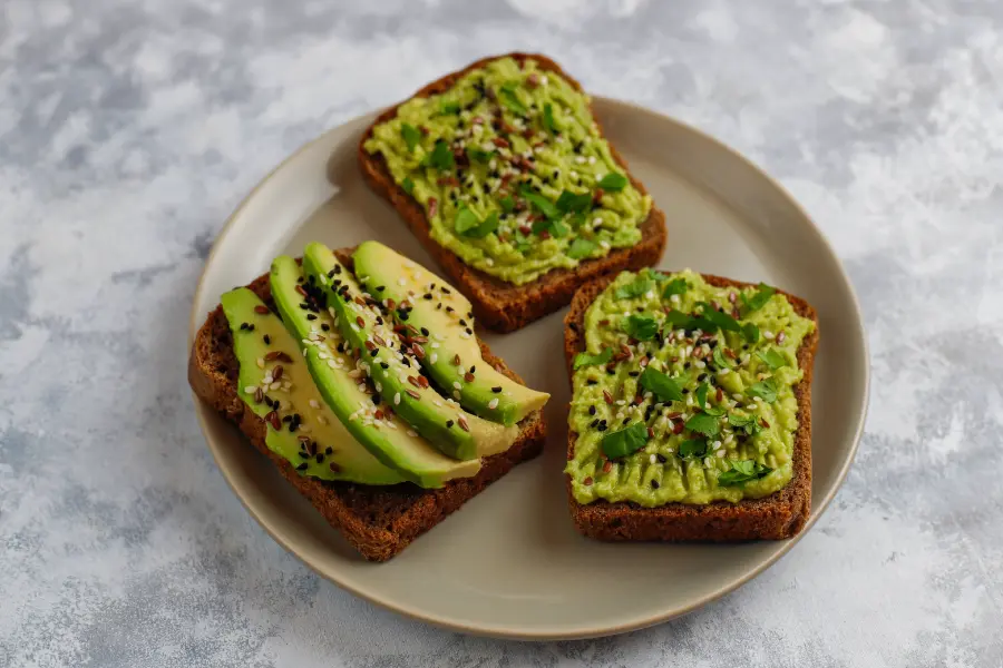 Open avocado toast with mashed avocado, sliced avocado, sesame seeds, flax seeds, and fresh herbs on dark bread, served on a beige plate.