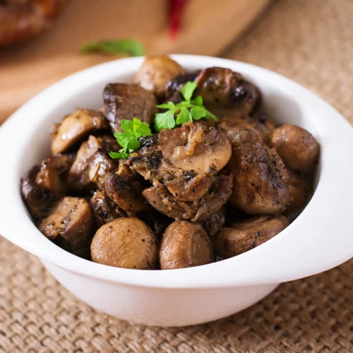 A bowl of baked mushrooms seasoned with herbs and garnished with fresh parsley, served in a white dish on a rustic burlap background.