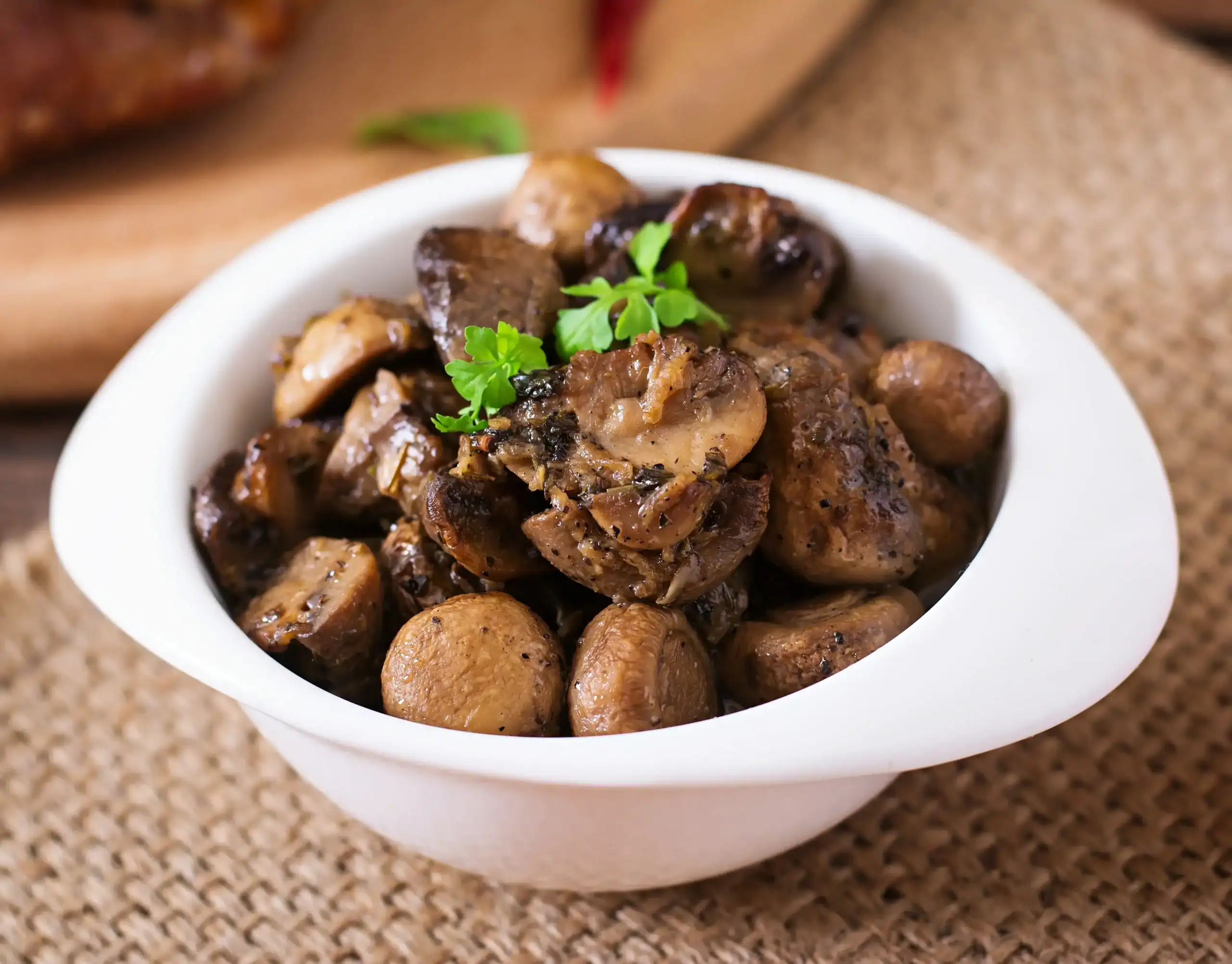 A bowl of baked mushrooms seasoned with herbs and garnished with fresh parsley, served in a white dish on a rustic burlap background.