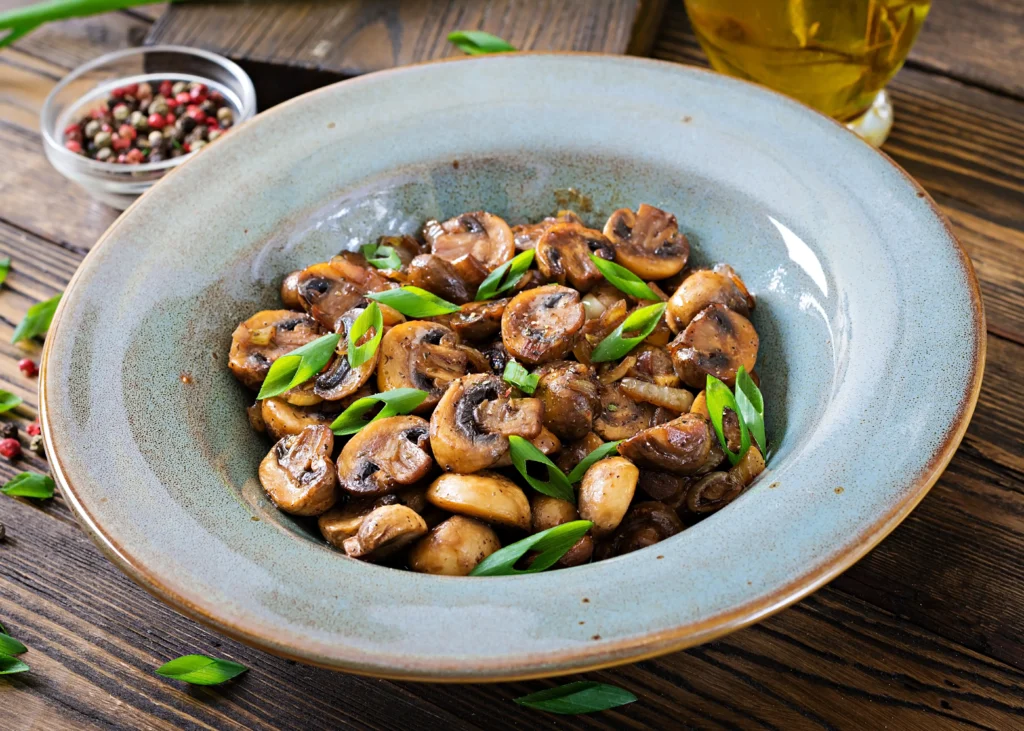 A rustic bowl filled with baked mushrooms, garnished with fresh green onions, served on a wooden table with peppercorns and olive oil in the background.