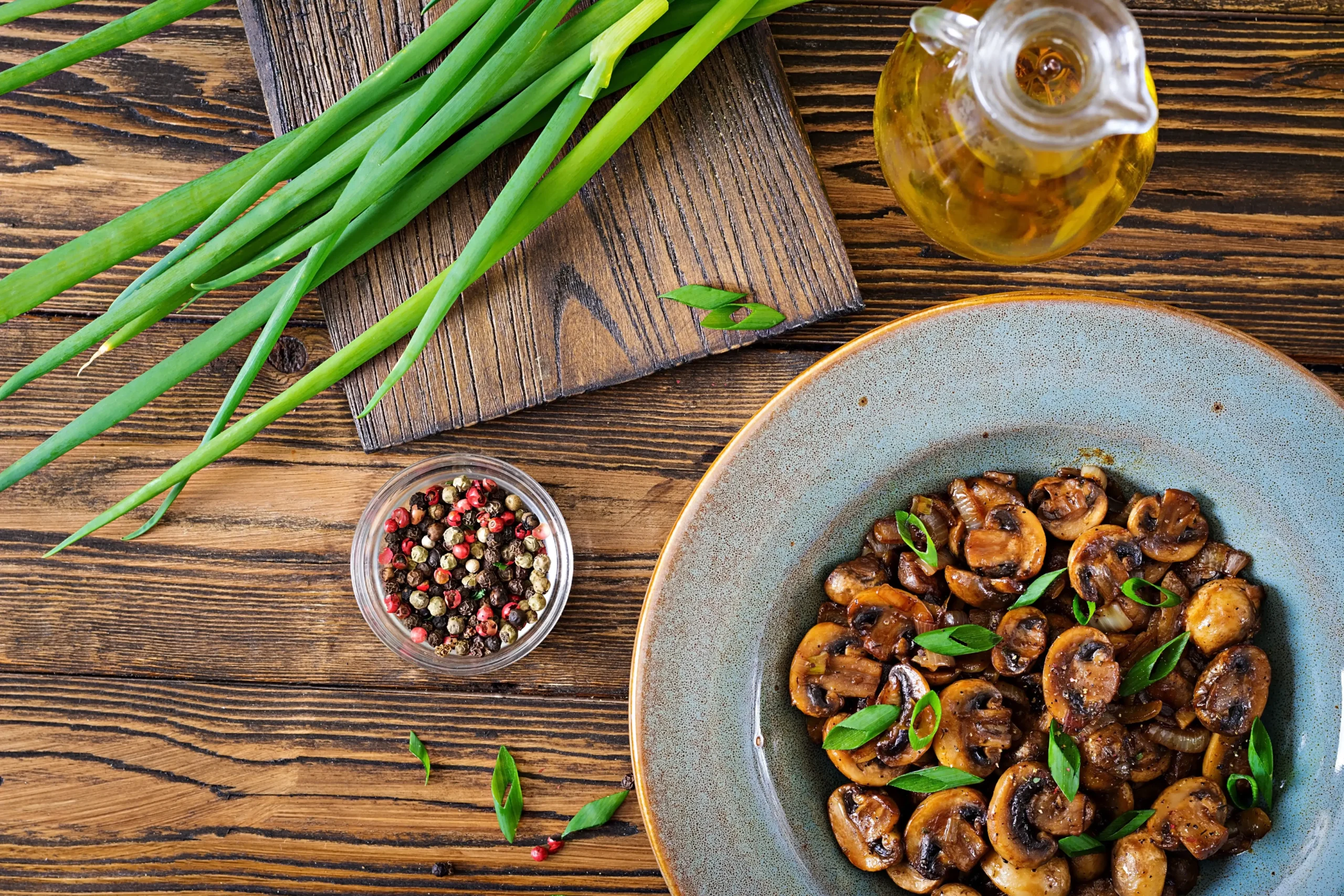 Top view of baked mushrooms garnished with green onions in a rustic bowl, accompanied by fresh scallions, colorful peppercorns, and olive oil on a wooden table.