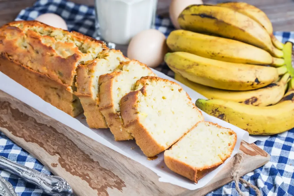 A freshly baked banana bread loaf sliced and displayed on a wooden cutting board, surrounded by ripe bananas, eggs, and a glass of milk.