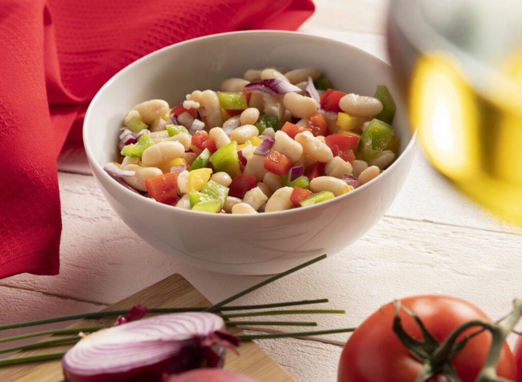 A fresh white bean salad with vibrant red and green bell peppers, diced tomatoes, and red onions, served in a white bowl with a red napkin in the background.