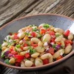 A colorful white bean salad with diced tomatoes, red onions, fresh herbs, and a drizzle of olive oil, served in a rustic ceramic bowl on a wooden table.