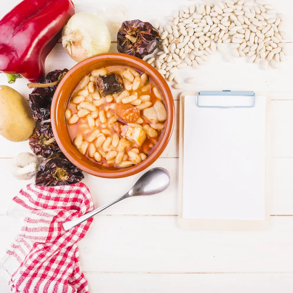 A bowl of hearty bean stew surrounded by fresh ingredients like a red bell pepper, onion, garlic, dried chili peppers, potatoes, and white beans, with a clipboard for notes on a white wooden background.