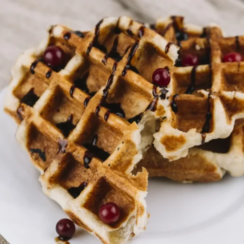 Close-up of golden Belgian waffles topped with chocolate syrup and red berries on a white plate.