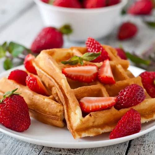 A plate of golden Belgian waffles topped with fresh strawberries and mint leaves, served on a rustic wooden table.