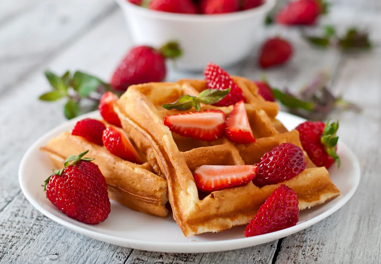 A plate of golden Belgian waffles topped with fresh strawberries and mint leaves, served on a rustic wooden table.