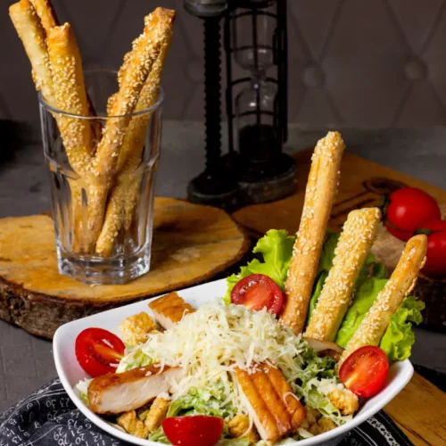 A fresh Chicken Caesar Salad served in a white bowl, topped with shredded cheese, grilled chicken, cherry tomatoes, croutons, and sesame breadsticks. A glass with more sesame breadsticks is placed beside the bowl, on a rustic wooden background.