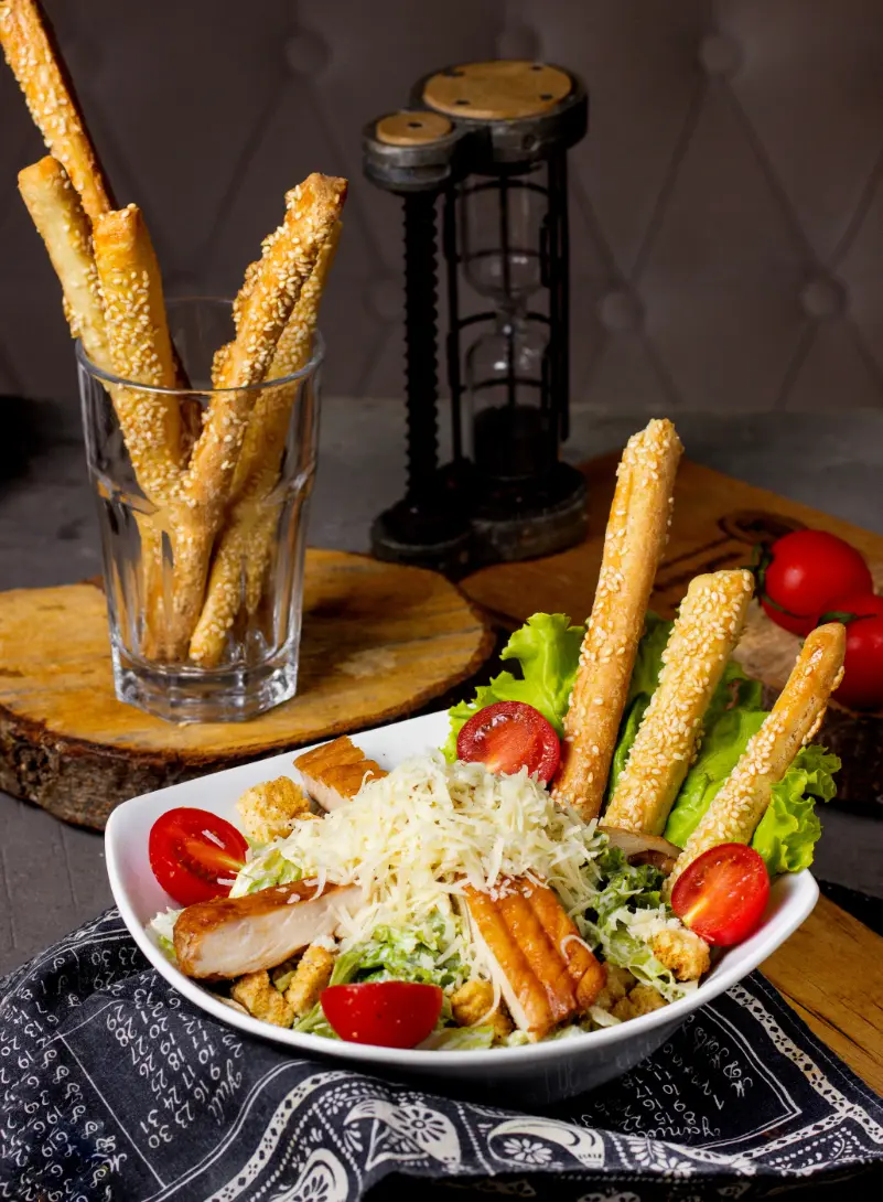 A fresh Chicken Caesar Salad served in a white bowl, topped with shredded cheese, grilled chicken, cherry tomatoes, croutons, and sesame breadsticks. A glass with more sesame breadsticks is placed beside the bowl, on a rustic wooden background.