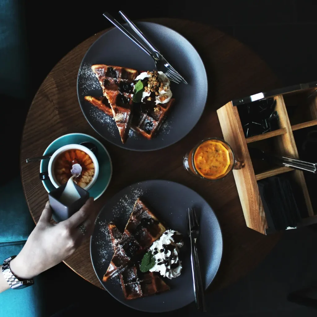 Overhead view of a table with two plates of beautifully presented waffles, topped with whipped cream, chocolate chips, and mint leaves, accompanied by coffee and orange juice.