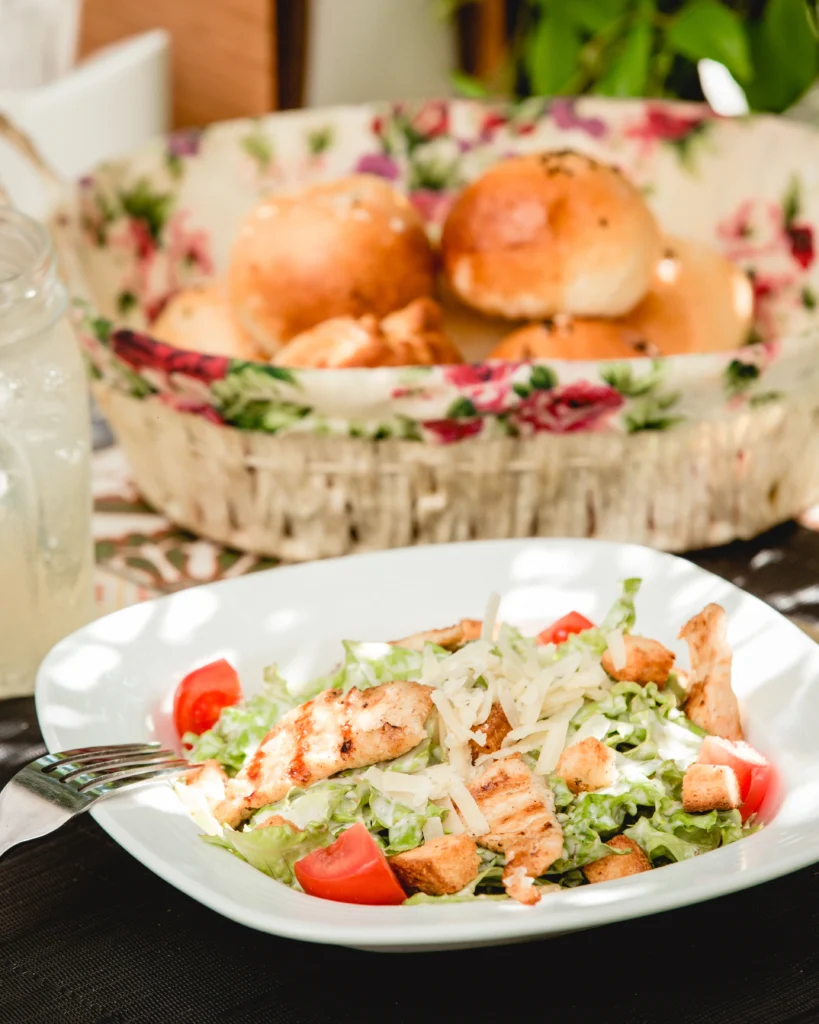 A vibrant Chicken Caesar Salad served on a white plate, featuring grilled chicken strips, shredded cheese, cherry tomatoes, lettuce, and crunchy croutons. In the background, a floral basket holds fresh dinner rolls.