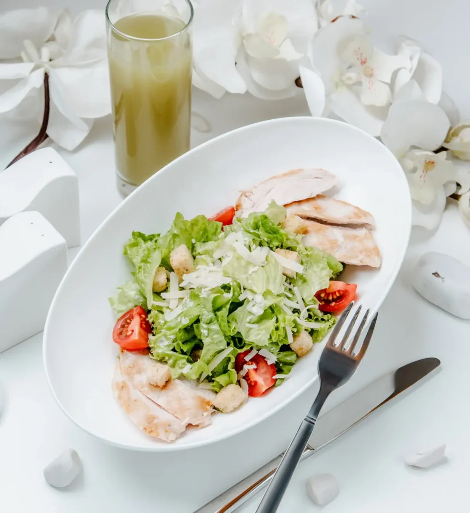 A pristine Chicken Caesar Salad served in an oval white dish, featuring grilled chicken slices, crisp lettuce, cherry tomatoes, croutons, and shredded cheese, accompanied by a glass of green juice and decorative white flowers in the background.