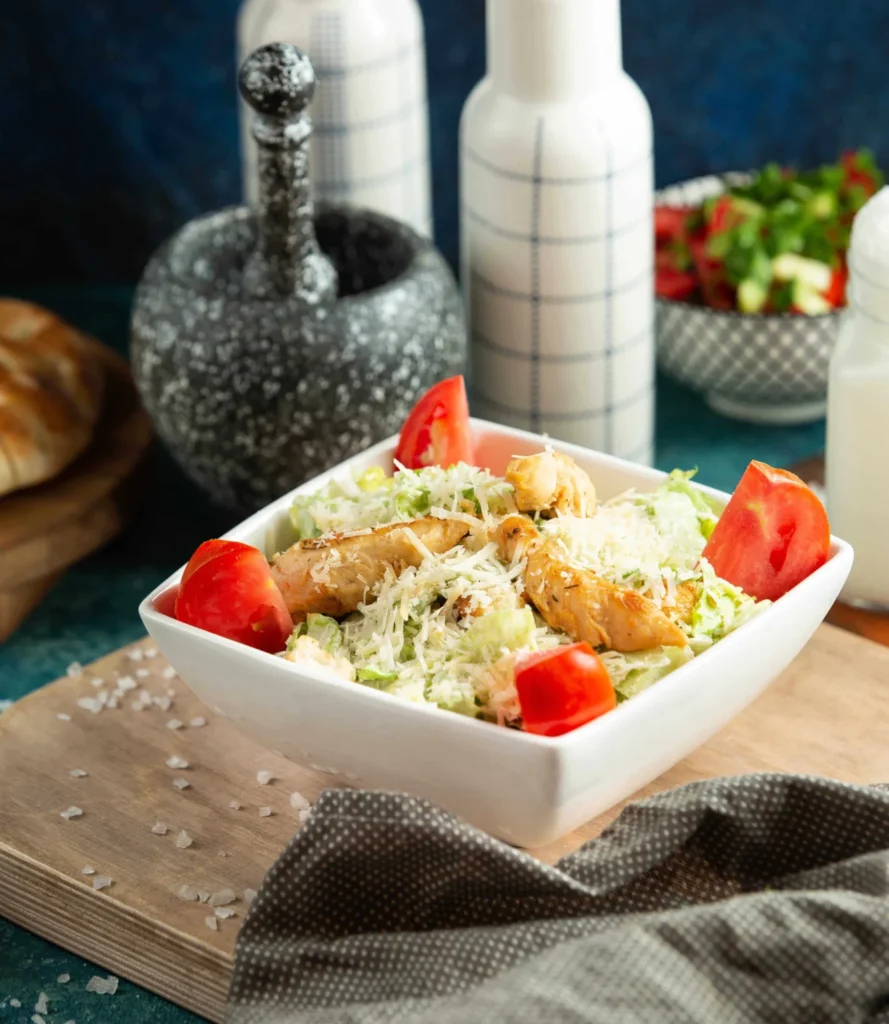 A bowl of chicken Caesar salad topped with shredded parmesan cheese, lettuce, grilled chicken, and tomato wedges, set on a rustic wooden board.