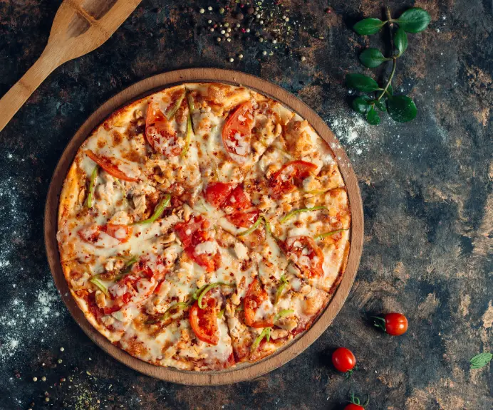 A delicious pizza topped with fresh tomatoes, bell peppers, melted cheese, and chicken, served on a round wooden board with scattered herbs and cherry tomatoes on a rustic surface.