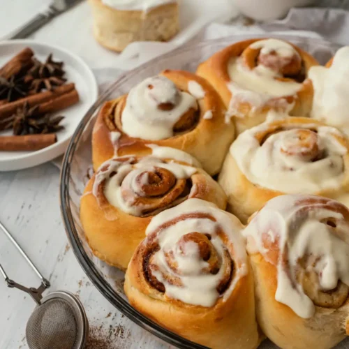 Homemade cinnamon rolls topped with cream cheese frosting in a glass baking dish, with cinnamon sticks and spices in the background.