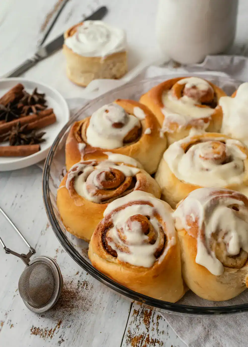 Homemade cinnamon rolls topped with cream cheese frosting in a glass baking dish, with cinnamon sticks and spices in the background.