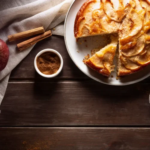 A beautifully baked apple pie with golden-brown crust, sliced and served on a white plate. Fresh apples, cinnamon sticks, and a bowl of cinnamon powder rest beside it on a rustic wooden table.