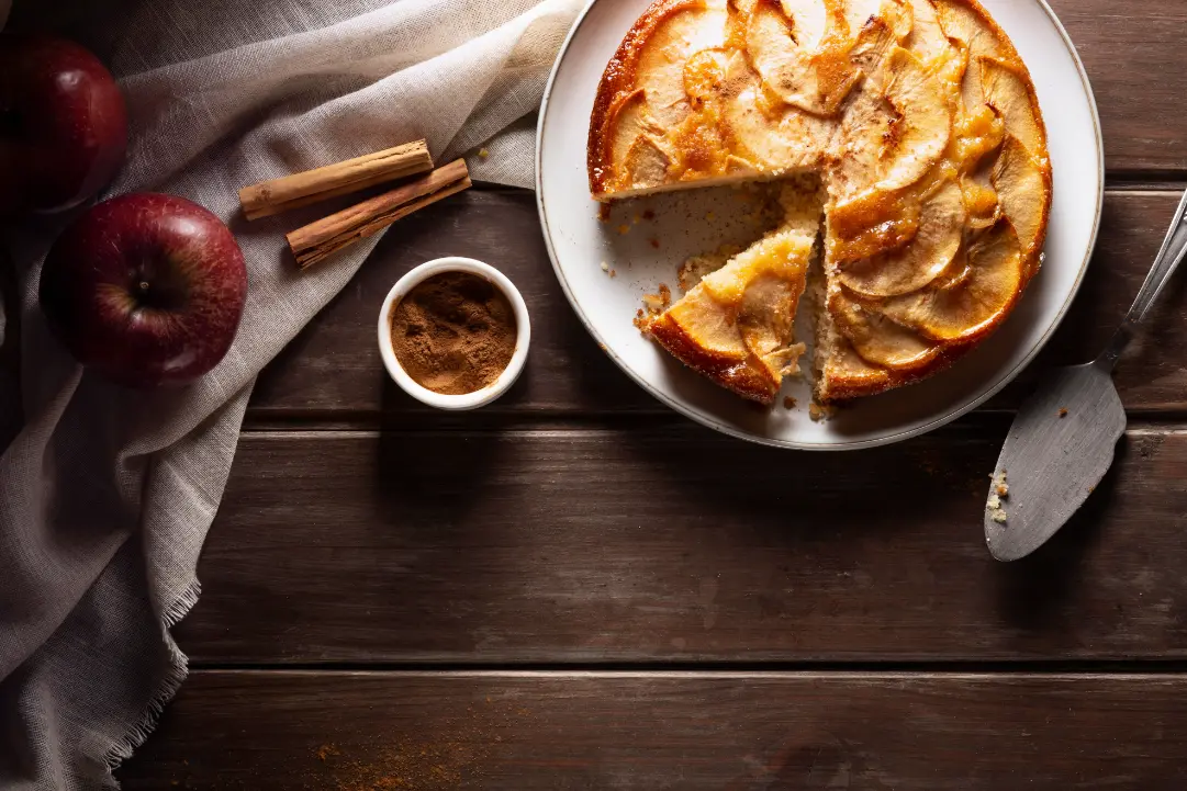 A beautifully baked apple pie with golden-brown crust, sliced and served on a white plate. Fresh apples, cinnamon sticks, and a bowl of cinnamon powder rest beside it on a rustic wooden table.