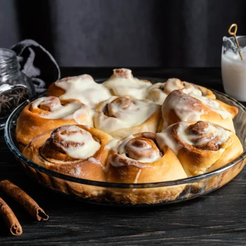 Freshly baked cinnamon rolls topped with cream cheese frosting in a glass baking dish, surrounded by cinnamon sticks and a glass of milk.