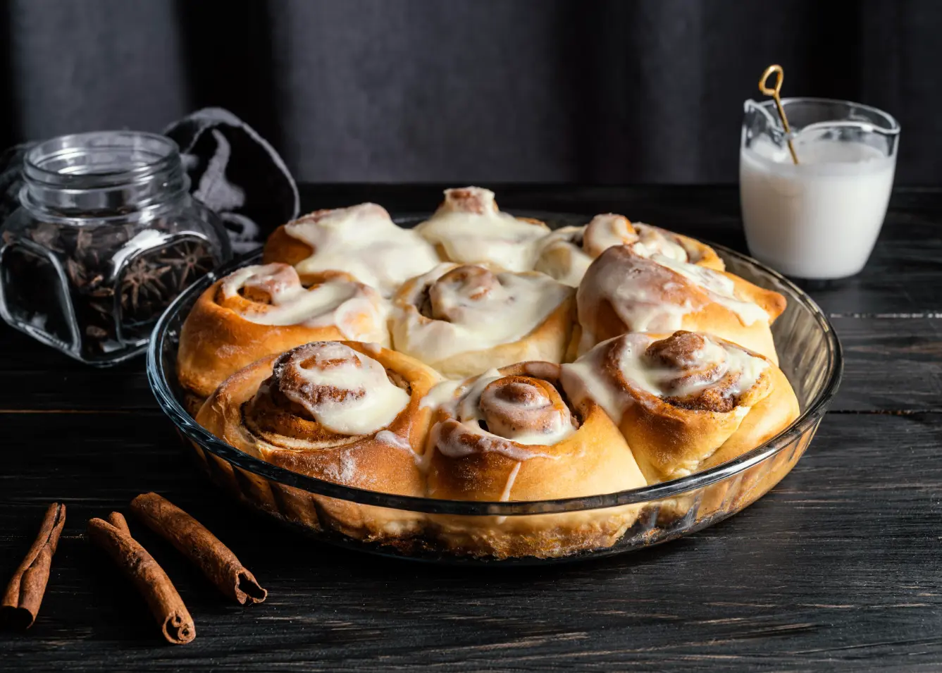 Freshly baked cinnamon rolls topped with cream cheese frosting in a glass baking dish, surrounded by cinnamon sticks and a glass of milk.