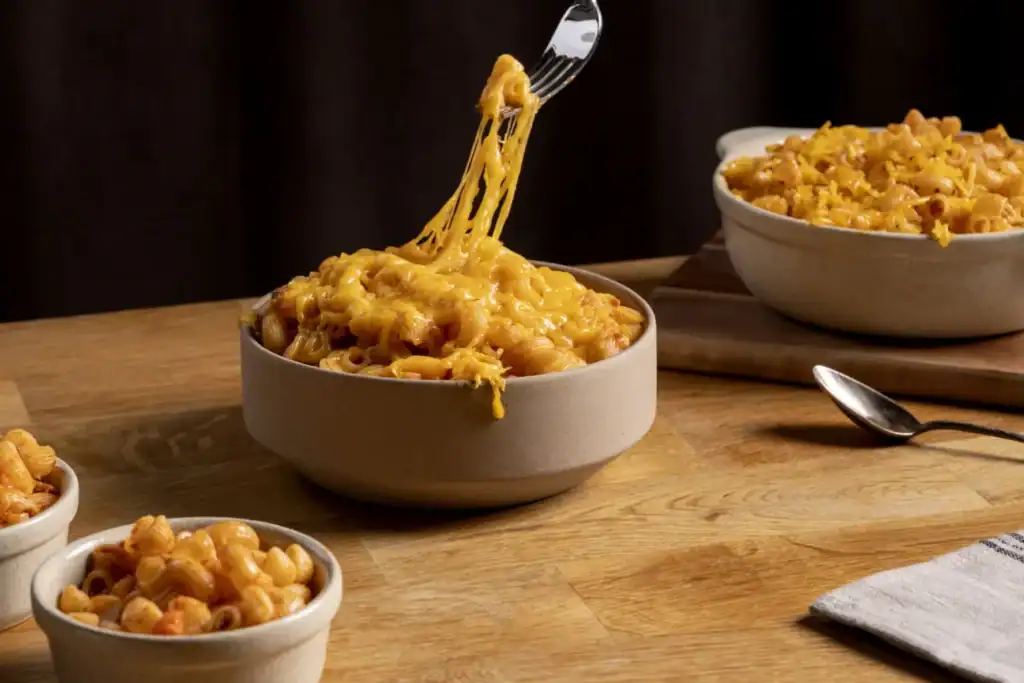 A bowl of creamy baked mac and cheese with a fork lifting a gooey, cheesy bite, surrounded by additional bowls of pasta on a wooden table.