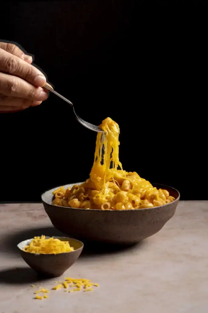 A bowl of baked mac and cheese with a hand lifting a cheesy bite using a fork, against a dark background.