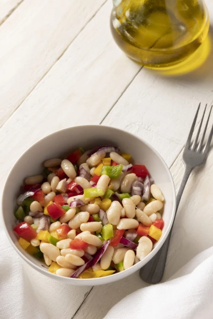 A colorful white bean salad with diced red and green bell peppers, red onions, and yellow peppers, served in a white bowl on a wooden table with a blank notepad for recipe notes.