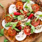 A vibrant veggie pizza topped with cherry tomatoes, sliced mushrooms, arugula, red chili peppers, pine nuts, and herbs, served on a wooden board with a "YUM!" flag decoration.
