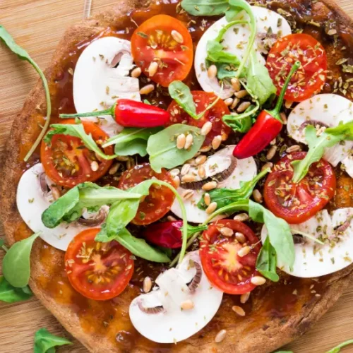 A vibrant veggie pizza topped with cherry tomatoes, sliced mushrooms, arugula, red chili peppers, pine nuts, and herbs, served on a wooden board with a "YUM!" flag decoration.