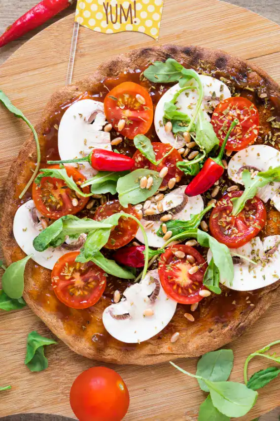 A vibrant veggie pizza topped with cherry tomatoes, sliced mushrooms, arugula, red chili peppers, pine nuts, and herbs, served on a wooden board with a "YUM!" flag decoration.