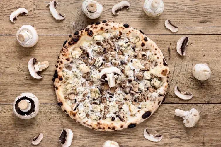 A freshly baked mushroom pizza topped with a variety of mushrooms and creamy cheese, surrounded by whole and sliced mushrooms on a wooden table.