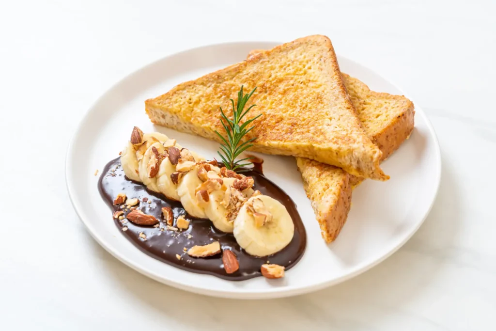 French toast served with banana slices, chocolate sauce, and crushed almonds on a white plate.