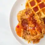 Close-up of crispy fried chicken drizzled with spicy syrup, served alongside a golden waffle on a white plate.