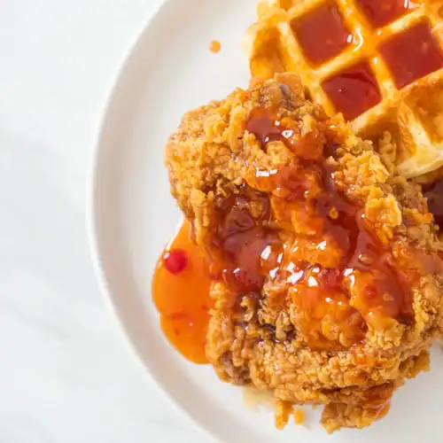 Close-up of crispy fried chicken drizzled with spicy syrup, served alongside a golden waffle on a white plate.