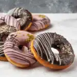 Chocolate and pink glazed doughnuts with decorative drizzles and toppings on a marble surface.