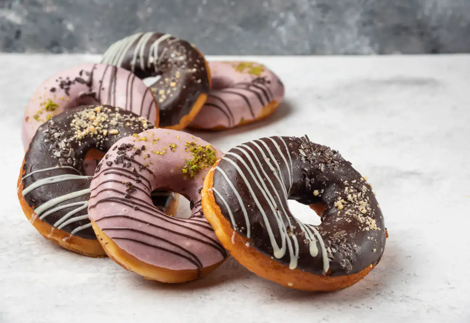 Chocolate and pink glazed doughnuts with decorative drizzles and toppings on a marble surface.
