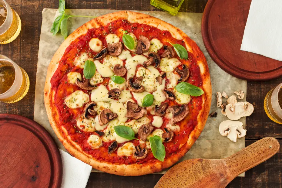 A freshly baked mushroom pizza topped with melted mozzarella, sliced mushrooms, fresh basil leaves, and tomato sauce, served on parchment paper with drinks and wooden serving boards.