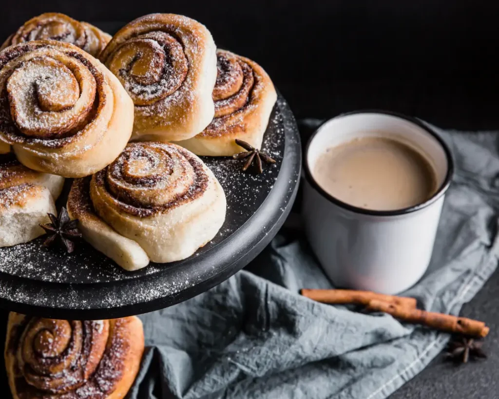 Freshly baked cinnamon rolls dusted with powdered sugar on a black cake stand, served with a cup of coffee.