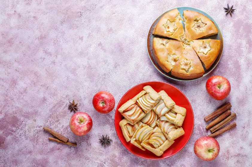 A beautifully styled image featuring a sliced apple pie with decorative crust, apple galettes on a red plate, and fresh apples, cinnamon sticks, and star anise scattered on a textured pink background.