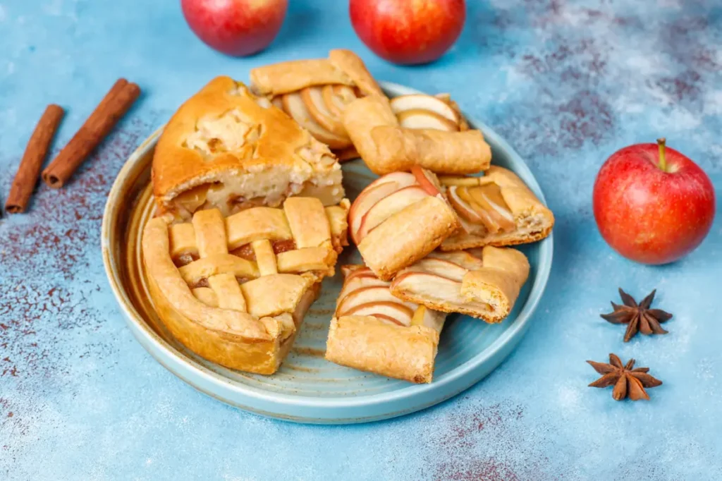 A beautifully styled image featuring a sliced apple pie with decorative crust, apple galettes on a red plate, and fresh apples, cinnamon sticks, and star anise scattered on a textured pink background.