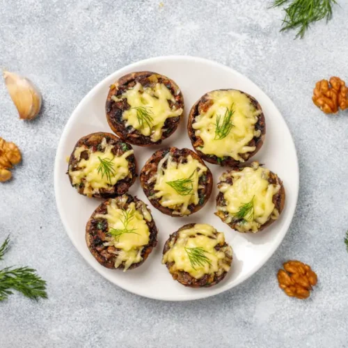 A plate of stuffed baked mushrooms garnished with melted cheese and fresh dill, surrounded by walnuts and garlic on a textured gray surface.