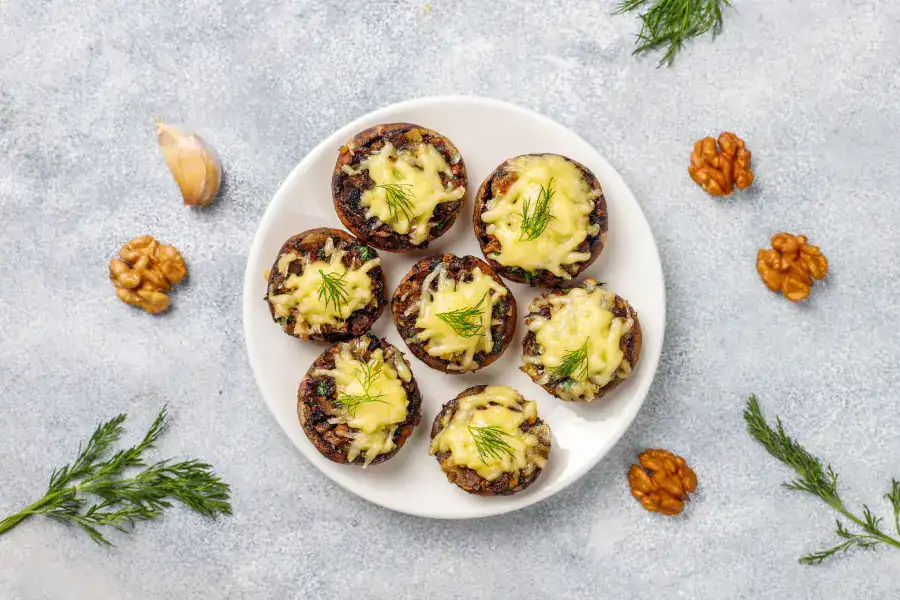 A plate of stuffed baked mushrooms garnished with melted cheese and fresh dill, surrounded by walnuts and garlic on a textured gray surface.